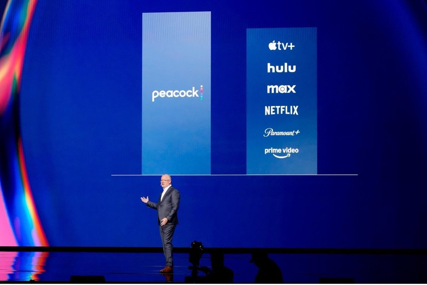 Man presents in front of a screen that shows Peacock and other streaming services.