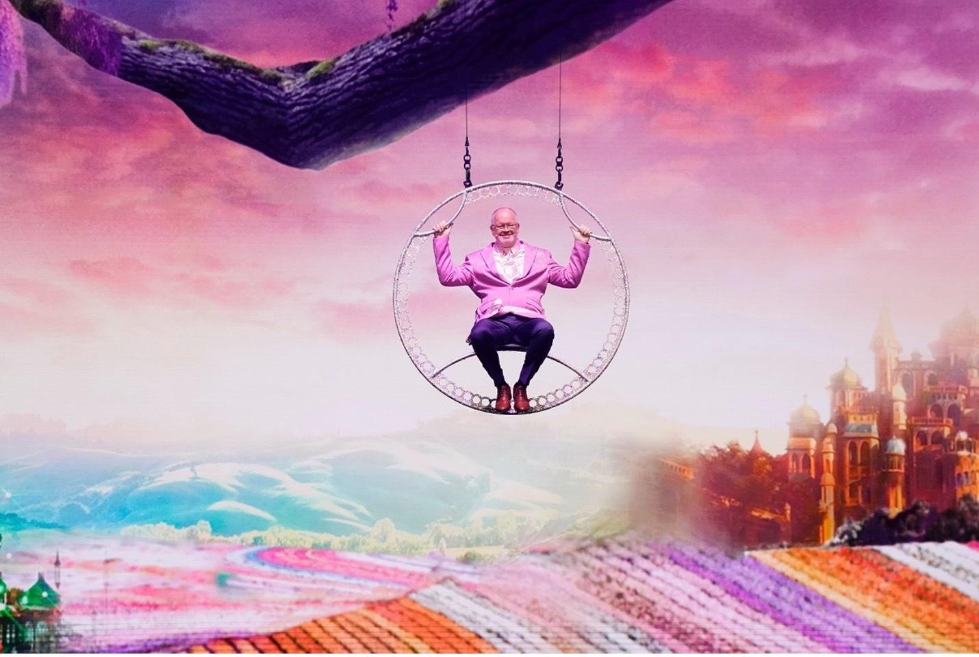 Man suspends from a circular fixture in front of a collage backdrop.