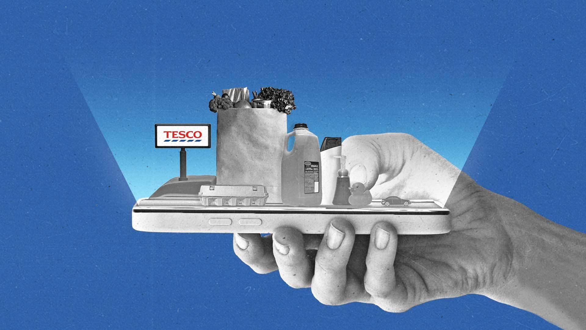 A hand holds a smartphone that features a checkout counter containing various items, including eggs, a shopping bag with groceries, a jug of orange juice, lotion, soap, a rubber duck and a toy car. A register at the end of the smartphone features a display showing the Tesco logo.