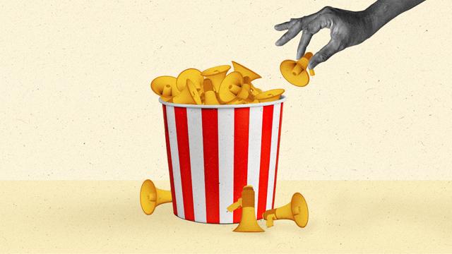 A hand removes a miniature yellow megaphone from a popcorn bucket filled with them.