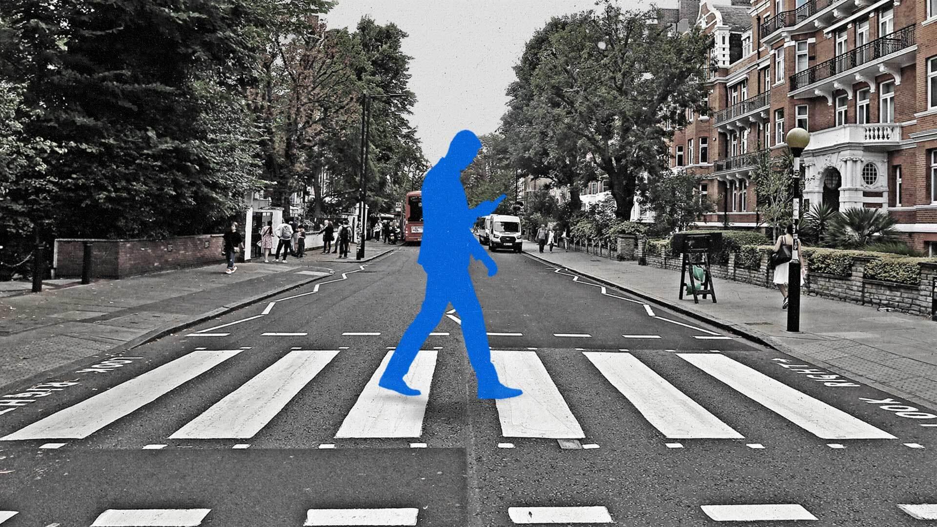 A man in blue crosses the street while looking at his smartphone.