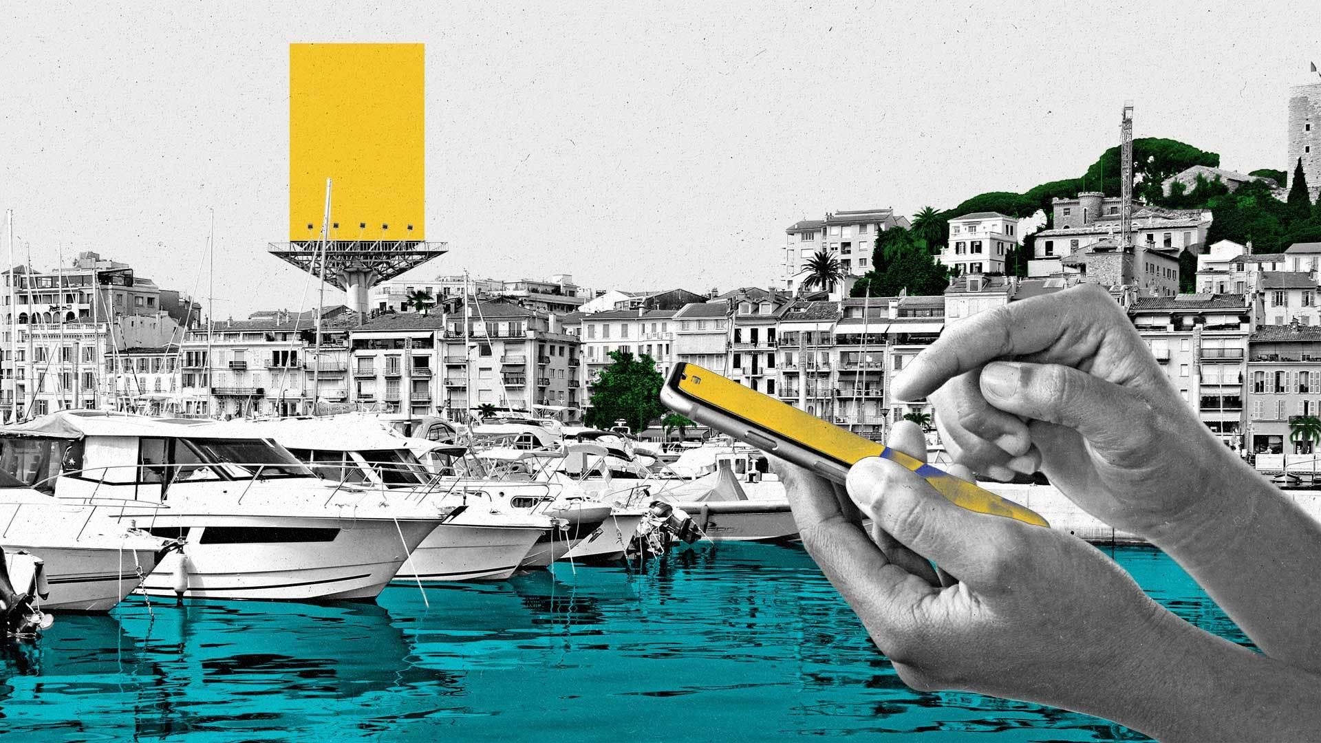 Harbor in Cannes with boats and buildings with a hand holding a mobile phone in the foreground.