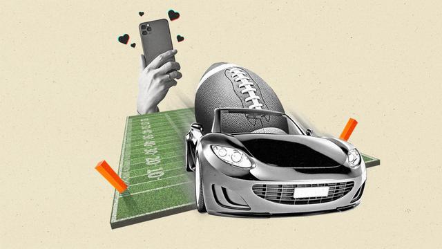 A car carrying an oversized football drives through the end zone of a football field as a hand holding a smartphone films it. Social media hearts float around the phone.